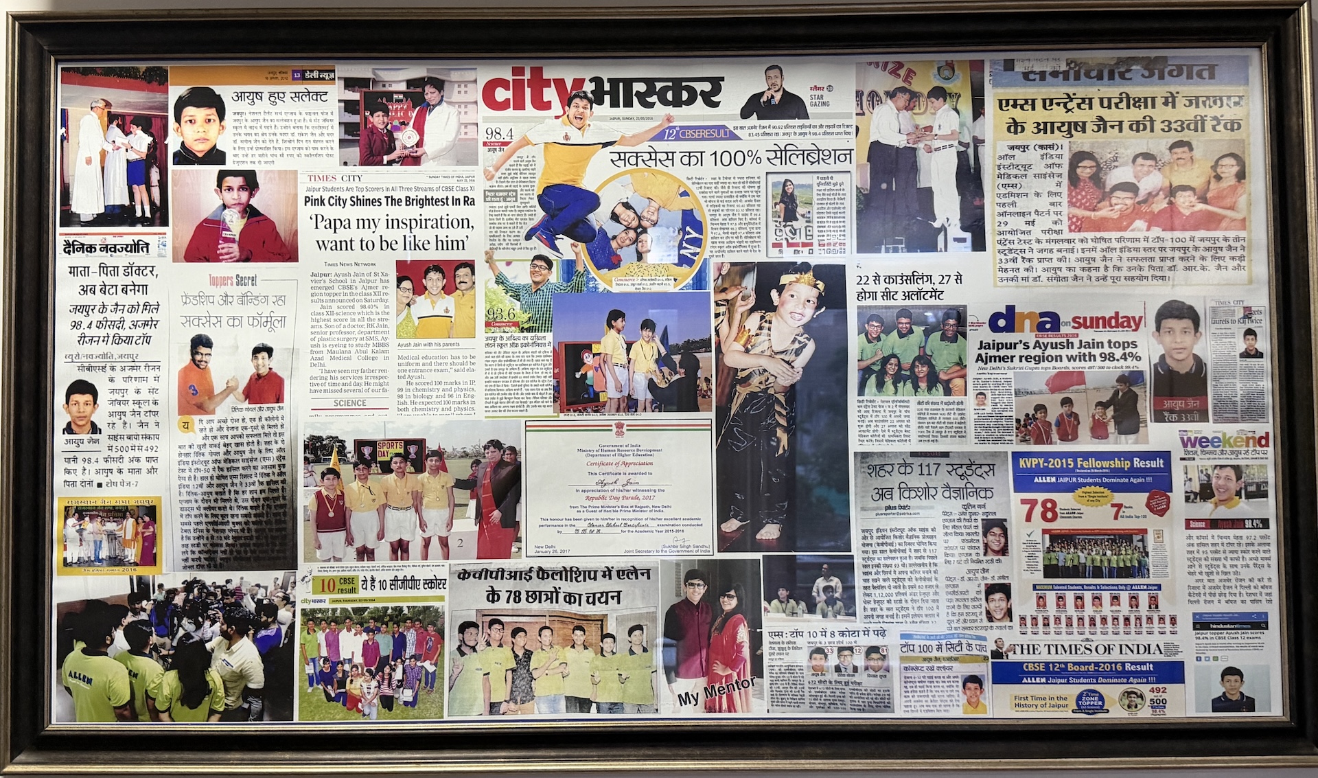 Framed collage of newspaper clippings covering Dr. Ayush Jain's academic achievements — CBSE Ajmer zonal topper, AIIMS entrance rank 33, KVPY fellowship and Republic Day parade invitation